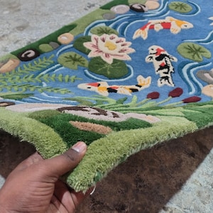 May include: A hand-tufted rug featuring a pond scene with koi fish, water lilies, and aquatic plants. The rug has a blue background with green, brown, and beige accents. The koi fish are orange, yellow, and black.