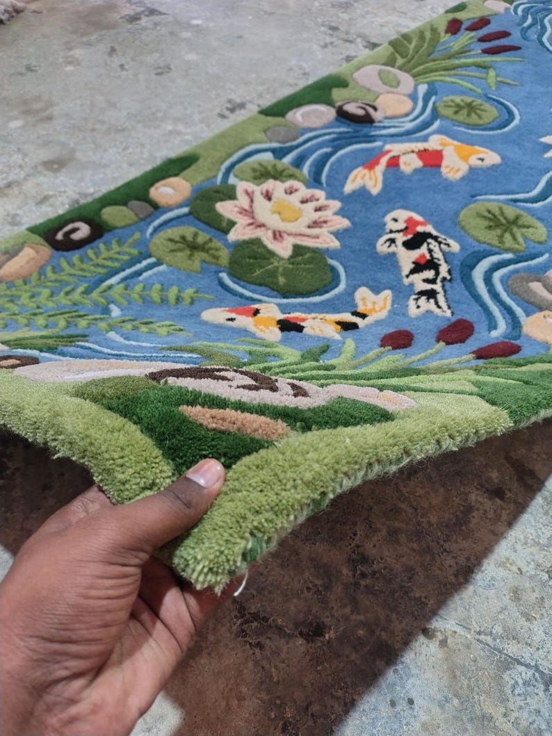 May include: A hand-tufted rug featuring a pond scene with koi fish, water lilies, and aquatic plants. The rug has a blue background with green, brown, and beige accents. The koi fish are orange, yellow, and black.