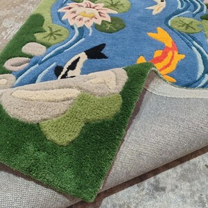 May include: A blue rug with a pond scene featuring koi fish, lily pads, and a lotus flower. The rug has green and beige accents, with a detailed design and a textured surface. The rug is folded over, revealing the backing.