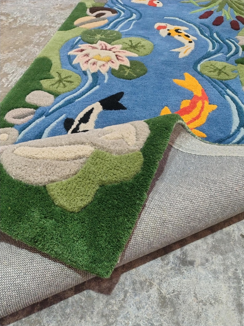 May include: A blue rug with a pond scene featuring koi fish, lily pads, and a lotus flower. The rug has green and beige accents, with a detailed design and a textured surface. The rug is folded over, revealing the backing.