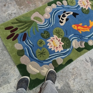 May include: A rectangular rug featuring a pond scene with a blue water background, green edges, and a variety of aquatic plants. Two koi fish, one black and white, the other orange, swim in the pond. The rug is on a concrete floor.