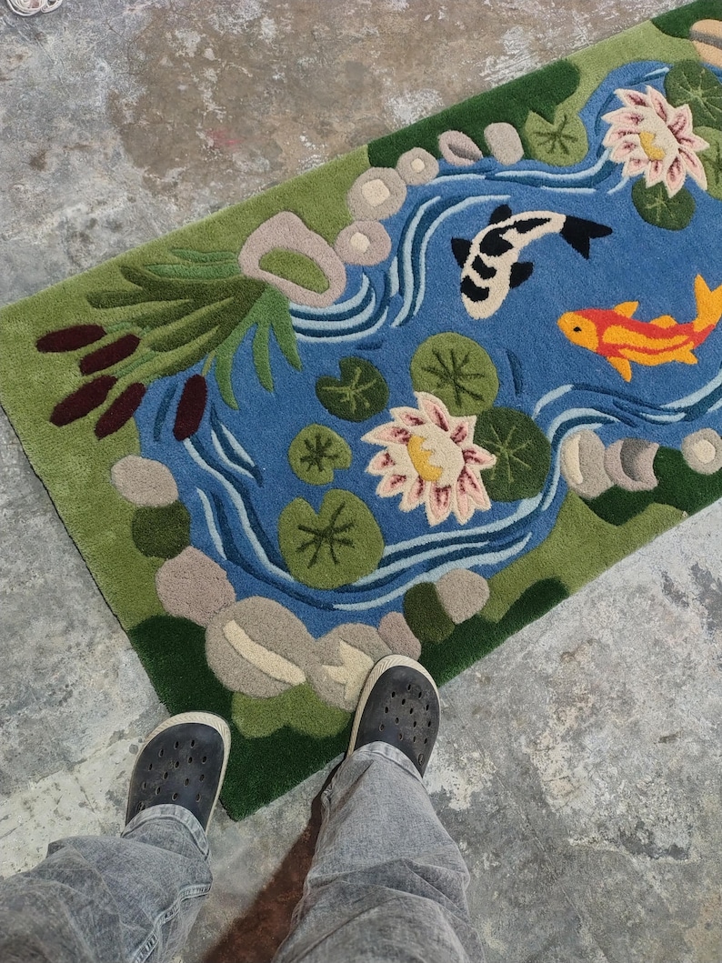 May include: A rectangular rug featuring a pond scene with a blue water background, green edges, and a variety of aquatic plants. Two koi fish, one black and white, the other orange, swim in the pond. The rug is on a concrete floor.