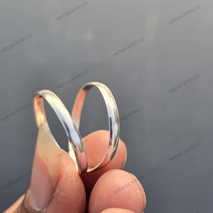 Puede incluir: Dos anillos de plata se muestran sobre un fondo gris. Los anillos tienen una superficie lisa y pulida, que refleja la luz. Son bandas sencillas, adecuadas para el uso diario o como anillos de boda. La imagen está bien iluminada, destacando el brillo metálico de los anillos.