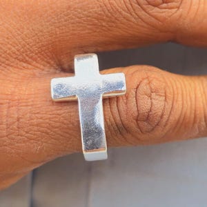 Pode incluir: Um anel prateado com um design de cruz. O anel, usado num dedo, tem um estilo minimalista. A cruz é o elemento central, com um acabamento polido. O design do anel é simples e elegante.