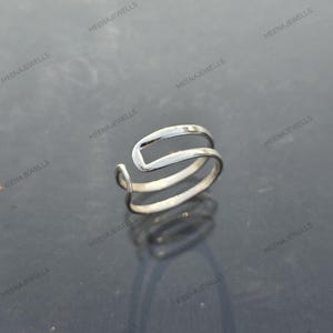Könnte beinhalten: Ein silberfarbener Ring mit einem einzigartigen Design. Der Ring hat eine gebogene, offene Form mit zwei Bändern. Der Markenname "MEENAJEWELLS" wiederholt sich um das Bild. Dieser Ring ist ein Schmuckstück.