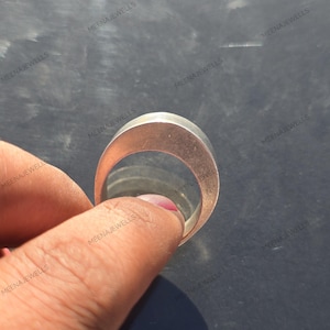 May include: A rose gold ring with a flat, wide band. The ring is held by a hand with a pink painted nail. The ring is smooth and shiny. The background is a dark, reflective surface. The brand name "MEENAJEWELLS" is watermarked on the image.