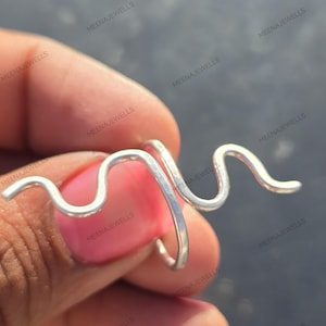 Könnte beinhalten: Ein silberfarbener Ring mit einem wellenförmigen Design. Der Ring hat eine einzigartige, abstrakte Form mit einer geschwungenen Linie, die sich um den Finger legt. Der Ring wird von einer Person mit rosa Nagellack gehalten. Der Markenname "MEENAJEWELLS" ist sichtbar.