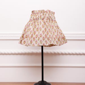 May include: A table lamp featuring a gathered fabric shade with a repeating pink and yellow floral pattern on a light beige background. The lamp has a slender black metal stem and a circular black base, providing a simple and functional design.
