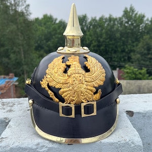 May include: A black helmet with a gold eagle emblem and a gold spike on top. The helmet has a leather brim and strap with gold-colored buckles. The background features green trees and a cloudy sky.