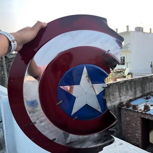 Peut inclure: Un bouclier de Captain America endommag&eacute;, avec un motif rouge, blanc et bleu et une &eacute;toile argent&eacute;e. Le bouclier pr&eacute;sente des signes d'usure, avec des dommages visibles sur les bords.