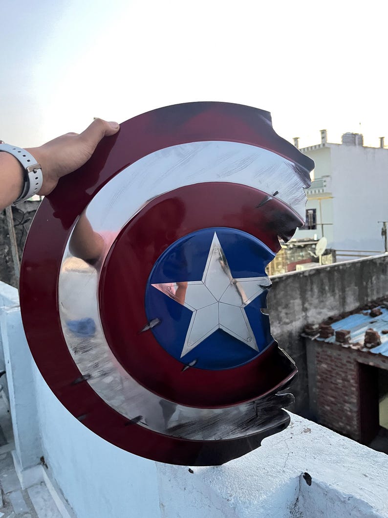 Peut inclure: Un bouclier de Captain America endommag&eacute;, avec un motif rouge, blanc et bleu et une &eacute;toile argent&eacute;e. Le bouclier pr&eacute;sente des signes d'usure, avec des dommages visibles sur les bords.