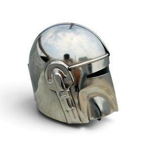 May include: A polished silver-colored helmet featuring a smooth, rounded dome, a horizontal visor opening, and a distinct T-shaped facial structure. The metallic surface reflects light, highlighting the curved contours and mechanical side details.