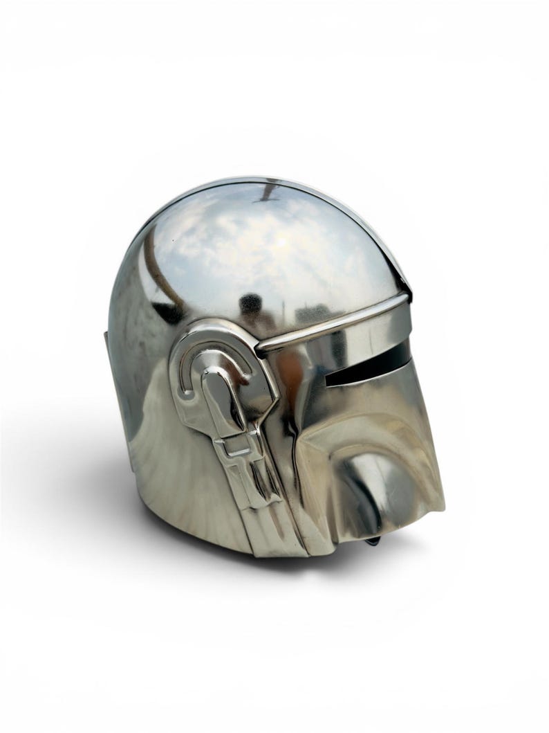 May include: A polished silver-colored helmet featuring a smooth, rounded dome, a horizontal visor opening, and a distinct T-shaped facial structure. The metallic surface reflects light, highlighting the curved contours and mechanical side details.
