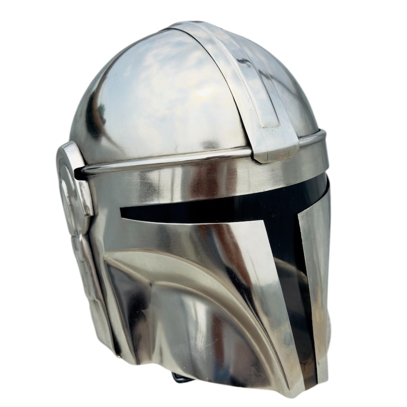 May include: A polished silver-colored metal helmet featuring a smooth, rounded dome, a prominent central ridge, and a T-shaped black visor opening. The design includes circular side ear covers and a contoured faceplate with a reflective finish.