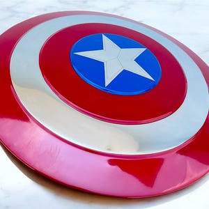 May include: A red, white, and blue shield with a central white star on a blue circle. The shield has a glossy finish and a circular design, reminiscent of a superhero's iconic accessory.