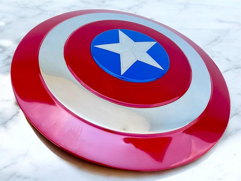 May include: A red, white, and blue shield with a central white star on a blue circle. The shield has a glossy finish and a circular design, reminiscent of a superhero's iconic accessory.