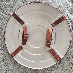 May include: A silver-colored, round shield with a red border and brown leather straps. The shield has a series of concentric circles and is designed with riveted metal accents. The straps are secured with buckles, and the shield appears to be a replica.