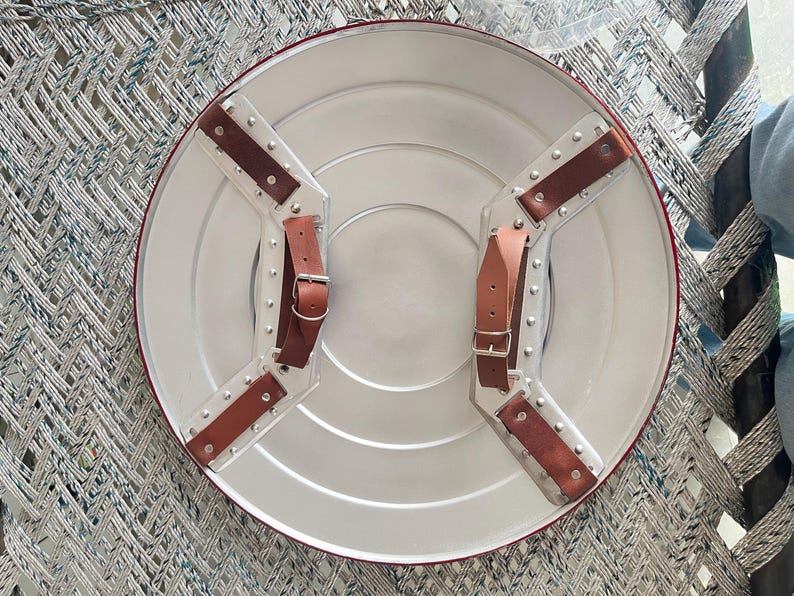 May include: A silver-colored, round shield with a red border and brown leather straps. The shield has a series of concentric circles and is designed with riveted metal accents. The straps are secured with buckles, and the shield appears to be a replica.