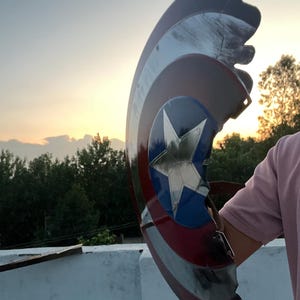 Peut inclure: Un bouclier endommag&eacute; de Captain America, avec une &eacute;toile bleue sur un cercle bleu, entour&eacute;e de rayures rouges et blanches. Le bouclier est pr&eacute;sent&eacute; devant un coucher de soleil, avec une partie manquante.