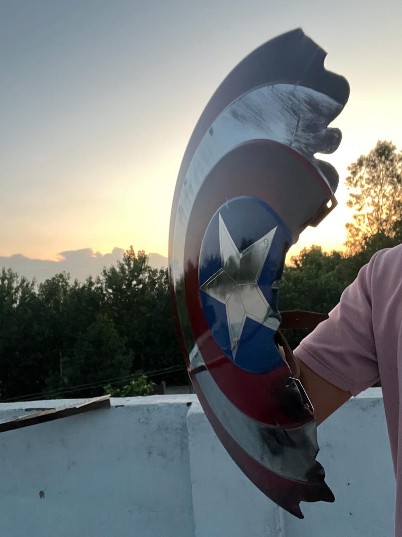Peut inclure: Un bouclier endommag&eacute; de Captain America, avec une &eacute;toile bleue sur un cercle bleu, entour&eacute;e de rayures rouges et blanches. Le bouclier est pr&eacute;sent&eacute; devant un coucher de soleil, avec une partie manquante.