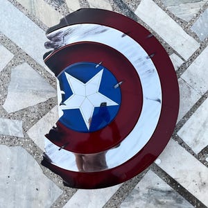 Peut inclure: Un bouclier Captain America vieilli, avec un sch&eacute;ma de couleurs rouge, blanc et bleu. Le bouclier pr&eacute;sente une &eacute;toile blanche sur un cercle bleu au centre, entour&eacute;e d'anneaux concentriques blancs et rouges. Le bouclier a une apparence us&eacute;e avec des bords &eacute;br&eacute;ch&eacute;s.