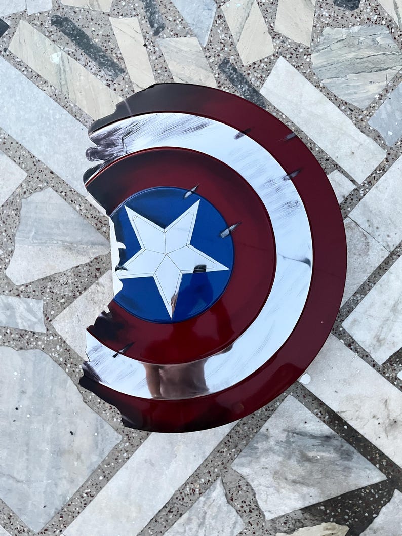 Peut inclure: Un bouclier Captain America vieilli, avec un sch&eacute;ma de couleurs rouge, blanc et bleu. Le bouclier pr&eacute;sente une &eacute;toile blanche sur un cercle bleu au centre, entour&eacute;e d'anneaux concentriques blancs et rouges. Le bouclier a une apparence us&eacute;e avec des bords &eacute;br&eacute;ch&eacute;s.