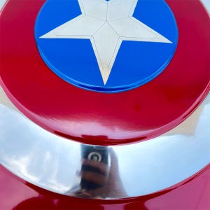 May include: A close-up of a Captain America shield. The shield features a red, white, and blue color scheme. The outer ring is red, with a silver-colored middle section, and a blue circle in the center with a white star.