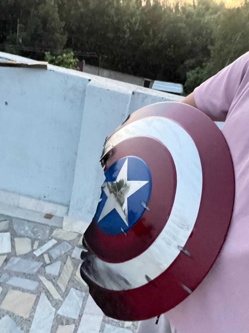 Peut inclure: Un bouclier de Captain America aux couleurs rouge, blanc et bleu. Le bouclier pr&eacute;sente une &eacute;toile blanche sur un cercle bleu, entour&eacute;e de bandes blanches et rouges concentriques. Le bouclier a une apparence us&eacute;e et endommag&eacute;e.