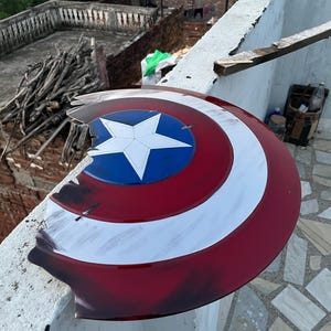 Peut inclure: Un bouclier Captain America endommag&eacute;, avec un sch&eacute;ma de couleurs rouge, blanc et bleu. Le bouclier pr&eacute;sente une &eacute;toile blanche sur un cercle bleu au centre, entour&eacute;e de rayures blanches et rouges. Le bouclier montre des signes de dommages.