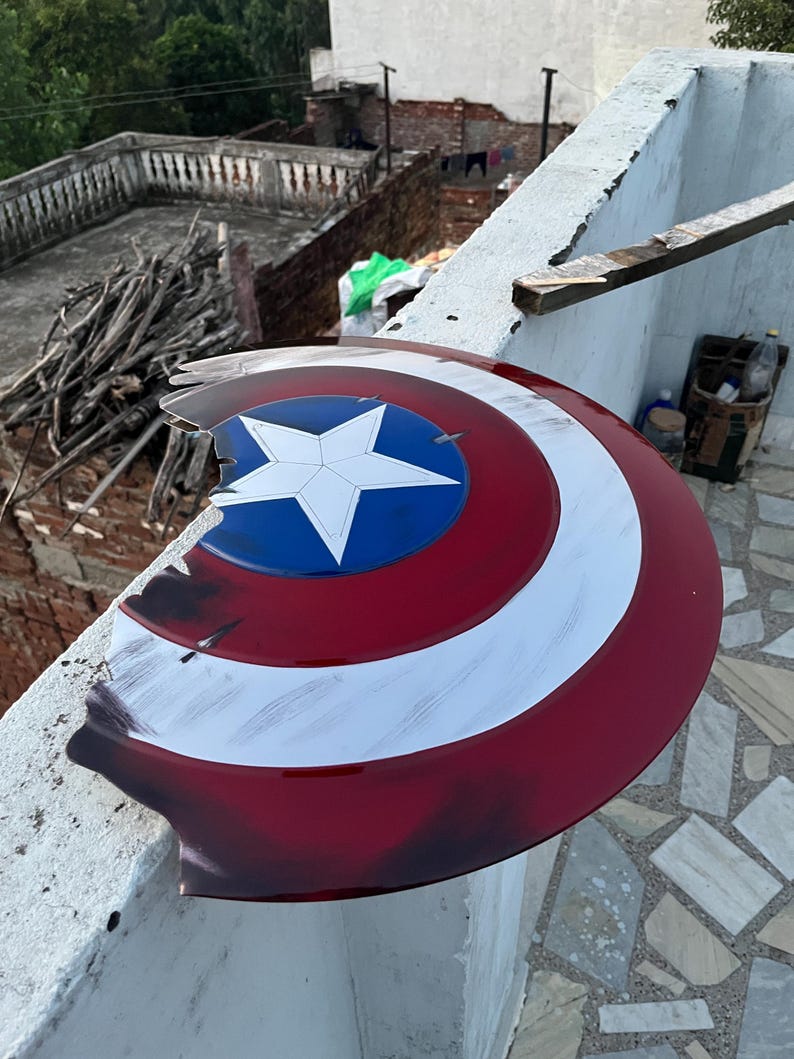 Peut inclure: Un bouclier Captain America endommag&eacute;, avec un sch&eacute;ma de couleurs rouge, blanc et bleu. Le bouclier pr&eacute;sente une &eacute;toile blanche sur un cercle bleu au centre, entour&eacute;e de rayures blanches et rouges. Le bouclier montre des signes de dommages.