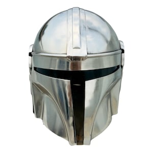 May include: A polished silver-colored metal helmet featuring a smooth, rounded dome, a central vertical ridge, and a horizontal T-shaped visor opening with a dark, reflective interior panel. The design includes contoured cheek plates and side details.