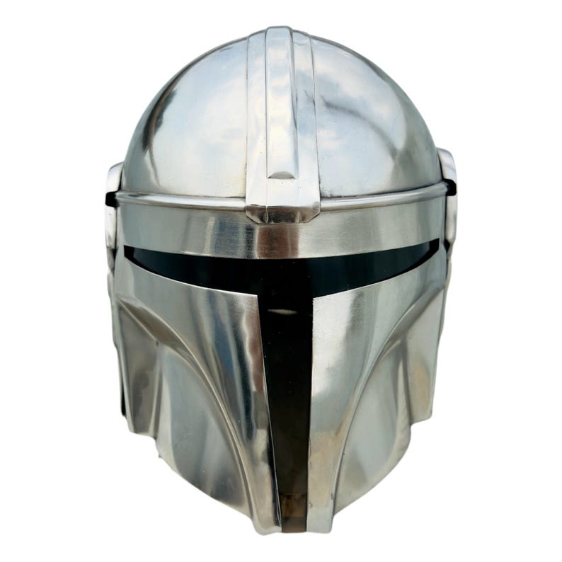 May include: A polished silver-colored metal helmet featuring a smooth, rounded dome, a central vertical ridge, and a horizontal T-shaped visor opening with a dark, reflective interior panel. The design includes contoured cheek plates and side details.