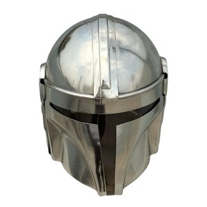 May include: A polished silver-colored metal helmet featuring a smooth, rounded dome, a central vertical ridge, and a horizontal visor opening. The design includes a T-shaped faceplate section with a dark, recessed area around the eye slit.