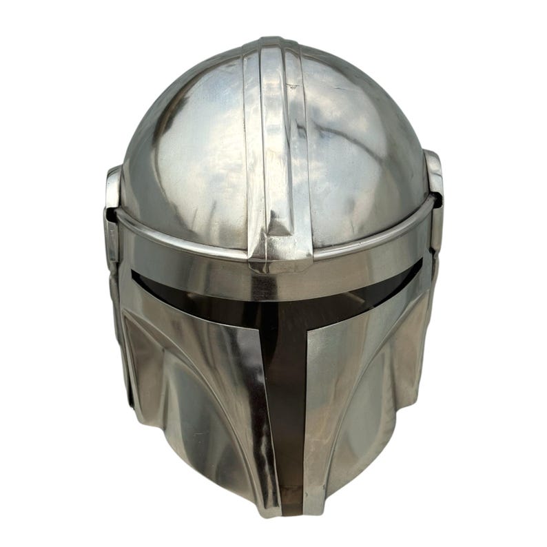 May include: A polished silver-colored metal helmet featuring a smooth, rounded dome, a central vertical ridge, and a horizontal visor opening. The design includes a T-shaped faceplate section with a dark, recessed area around the eye slit.