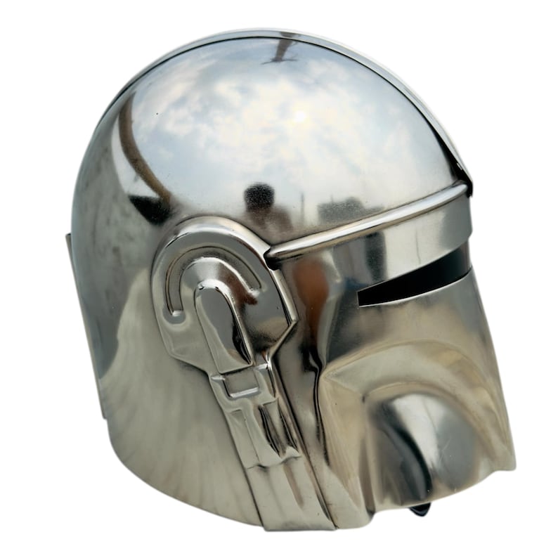 May include: A polished silver-colored metal helmet featuring a smooth, rounded dome, a horizontal visor opening, and a molded faceplate with integrated side ear-piece detailing. The reflective surface shows distorted environmental reflections.