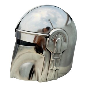 May include: A polished silver-colored metal helmet featuring a smooth, rounded dome, a horizontal visor opening, and a distinct T-shaped facial structure. The surface is highly reflective, showing light and environmental reflections across its curved exterior.