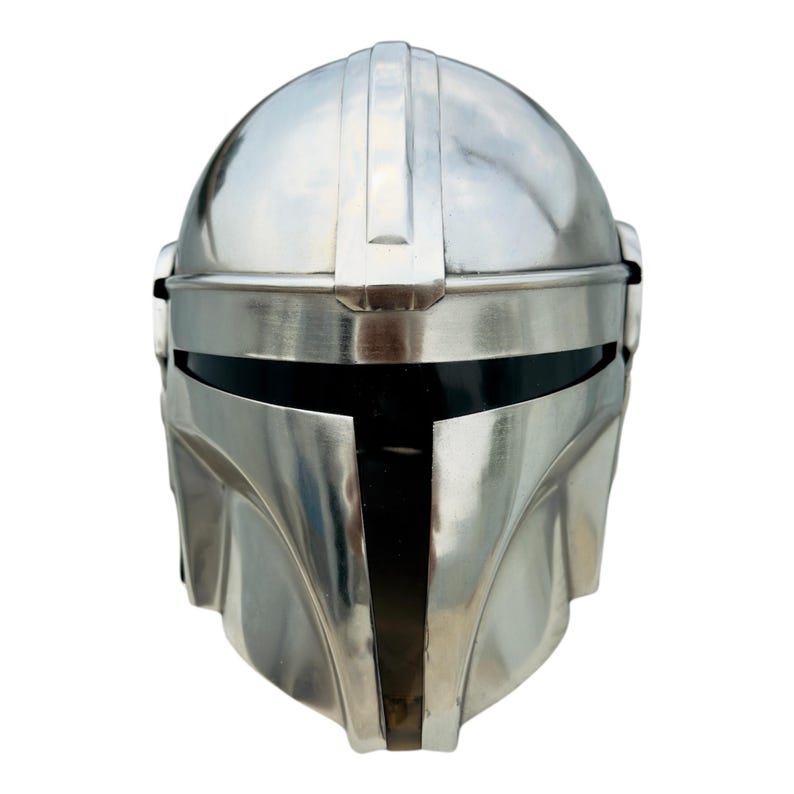 May include: A polished silver-colored metal helmet featuring a smooth, rounded dome, a central vertical ridge, and a horizontal T-shaped visor opening with a dark, recessed interior. The design includes contoured cheek plates and a curved lower edge.