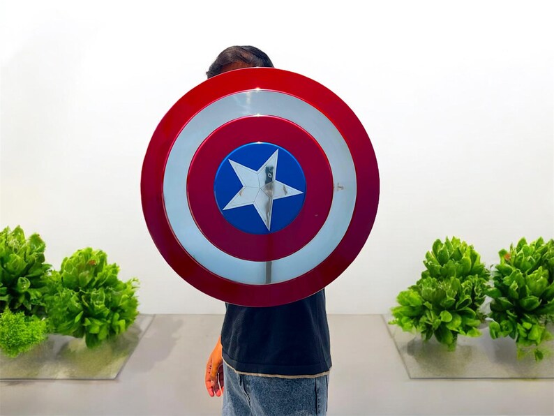 May include: A round shield with a red, white, and blue design, featuring a white star in the center. The shield is held in front of a person. The shield is a prop or costume accessory.
