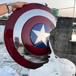 Peut inclure: Un bouclier Captain America, avec un sch&eacute;ma de couleurs rouge, blanc et bleu. Le bouclier pr&eacute;sente une &eacute;toile en son centre et un anneau argent&eacute; m&eacute;tallique. Le bouclier a un bord endommag&eacute;.
