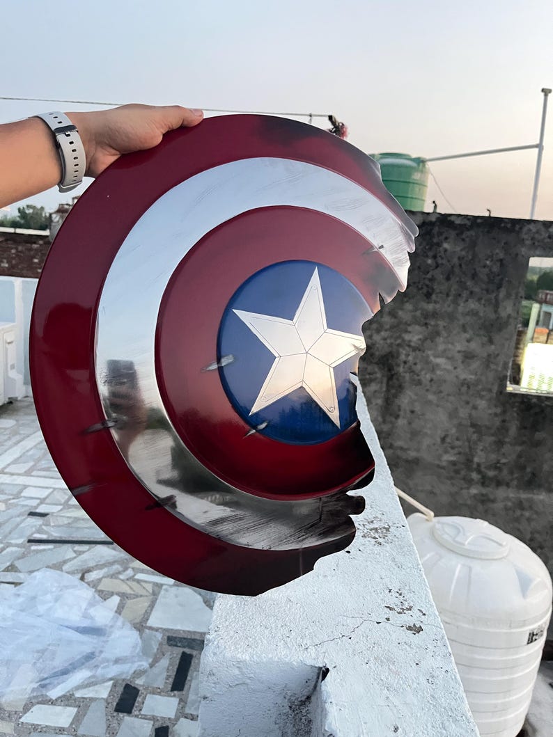 Peut inclure: Un bouclier Captain America, avec un sch&eacute;ma de couleurs rouge, blanc et bleu. Le bouclier pr&eacute;sente une &eacute;toile en son centre et un anneau argent&eacute; m&eacute;tallique. Le bouclier a un bord endommag&eacute;.