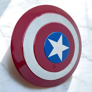 May include: A red, white, and blue shield with a star emblem. The shield has a glossy finish and a circular design, reminiscent of a superhero's iconic accessory. The shield is a prop or collectible.