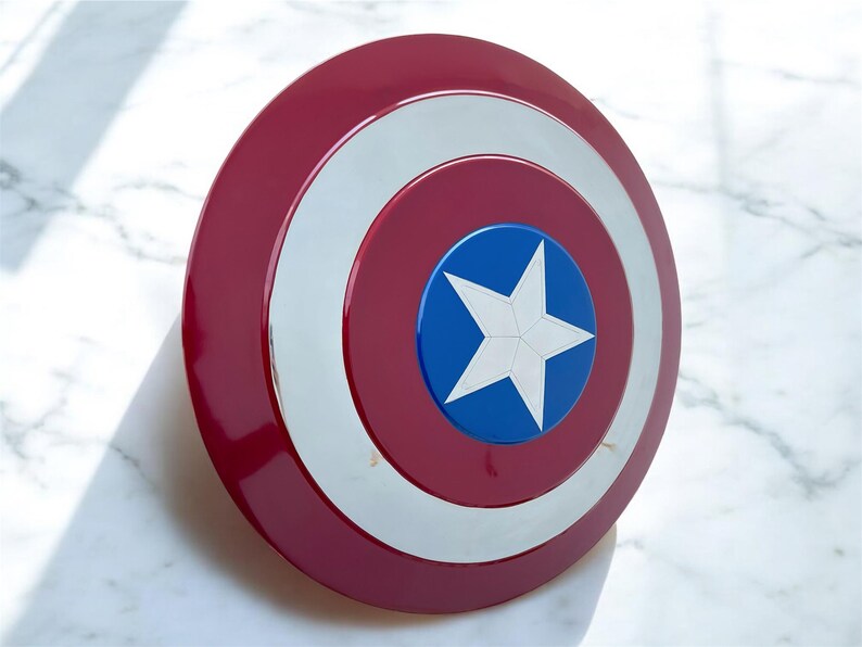 May include: A red, white, and blue shield with a star emblem. The shield has a glossy finish and a circular design, reminiscent of a superhero's iconic accessory. The shield is a prop or collectible.