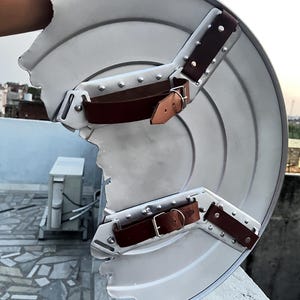 Peut inclure: Un bouclier argent&eacute; endommag&eacute;, avec des sangles en cuir marron et des boucles m&eacute;talliques. Le bouclier a un design circulaire avec une partie manquante sur le bord. Les sangles sont fix&eacute;es avec des rivets.