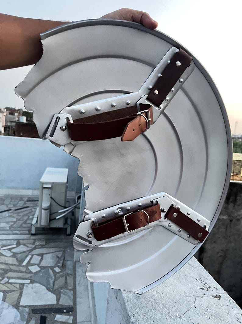Peut inclure: Un bouclier argent&eacute; endommag&eacute;, avec des sangles en cuir marron et des boucles m&eacute;talliques. Le bouclier a un design circulaire avec une partie manquante sur le bord. Les sangles sont fix&eacute;es avec des rivets.