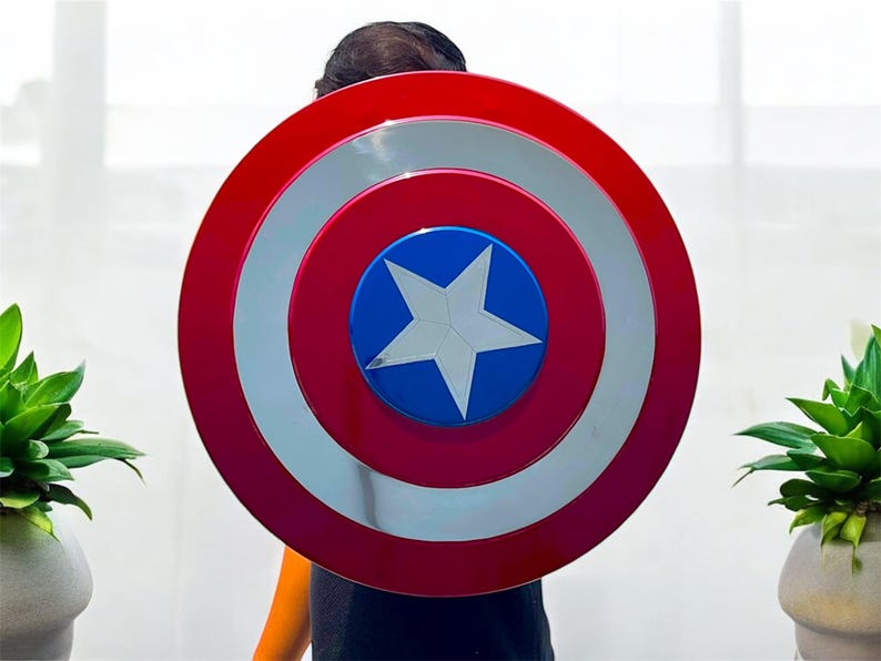 May include: A red, white, and blue shield with a star in the center. The shield has a circular design with concentric rings of red, silver, and red. The center is blue with a white star.