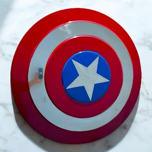 May include: A red, white, and blue shield with a star in the center. The shield has a circular design with concentric rings of red, white, and red. The center is blue with a white star.