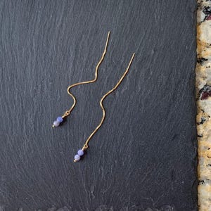 May include: A pair of gold-tone threader earrings featuring a delicate chain design. Each earring terminates in two small, round, light purple beads. The earrings are constructed with a thin, flexible metal chain that hangs in a gentle curve.