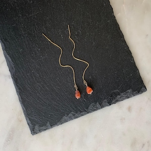 May include: A pair of gold-toned threader earrings featuring long, wavy metal chains. Each earring terminates in a small, polished, irregular-shaped orange-red gemstone bead that hangs from the end of the delicate gold chain.