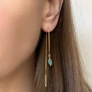 Puede incluir: Un pendiente de hilo dorado con una cadena delicada que atraviesa el lóbulo de la oreja. Un extremo de la cadena termina en una barra metálica recta, mientras que el otro extremo presenta una pequeña cuenta de piedra bruta de color verde claro.