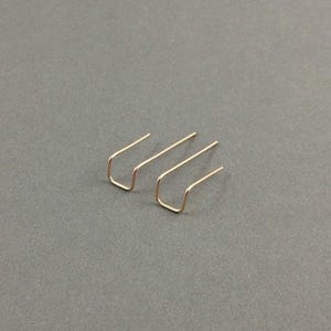 May include: A pair of rose gold-toned ear climbers, each featuring a simple, slender wire bent into a U-shape with two parallel prongs extending upward. The earrings have a smooth, polished metallic finish and a minimalist, open-frame design.